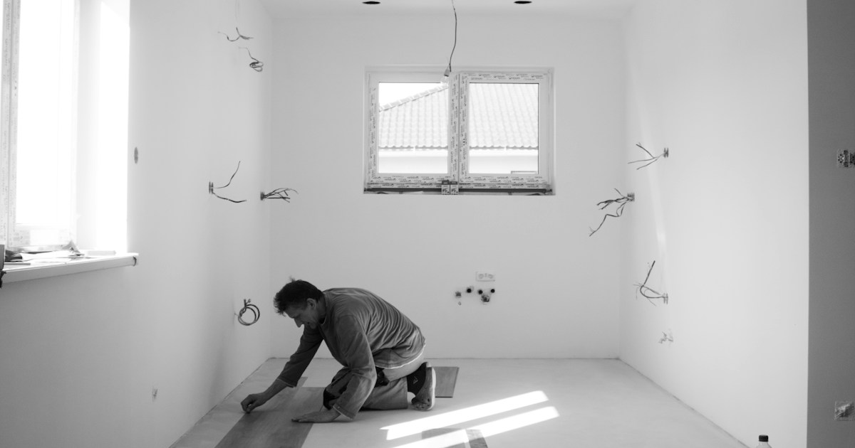 Flooring installer kneeling and laying hardwood planks in a room under construction
